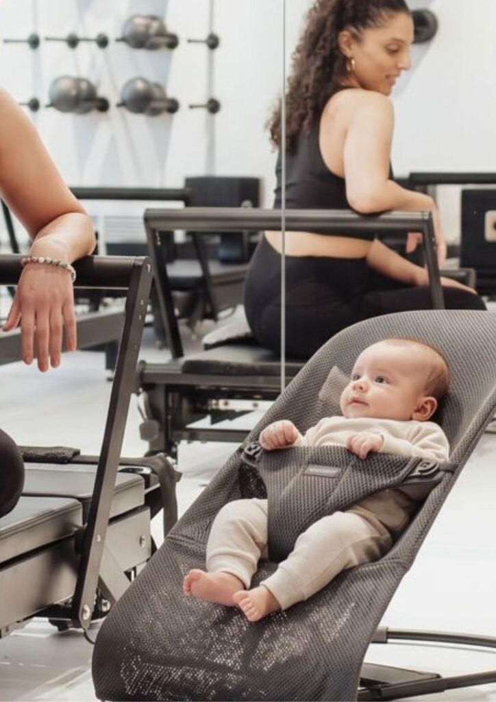 Baby in a BabyBjörn bouncer beside a Reformer during a postnatal Pilates class in Munich